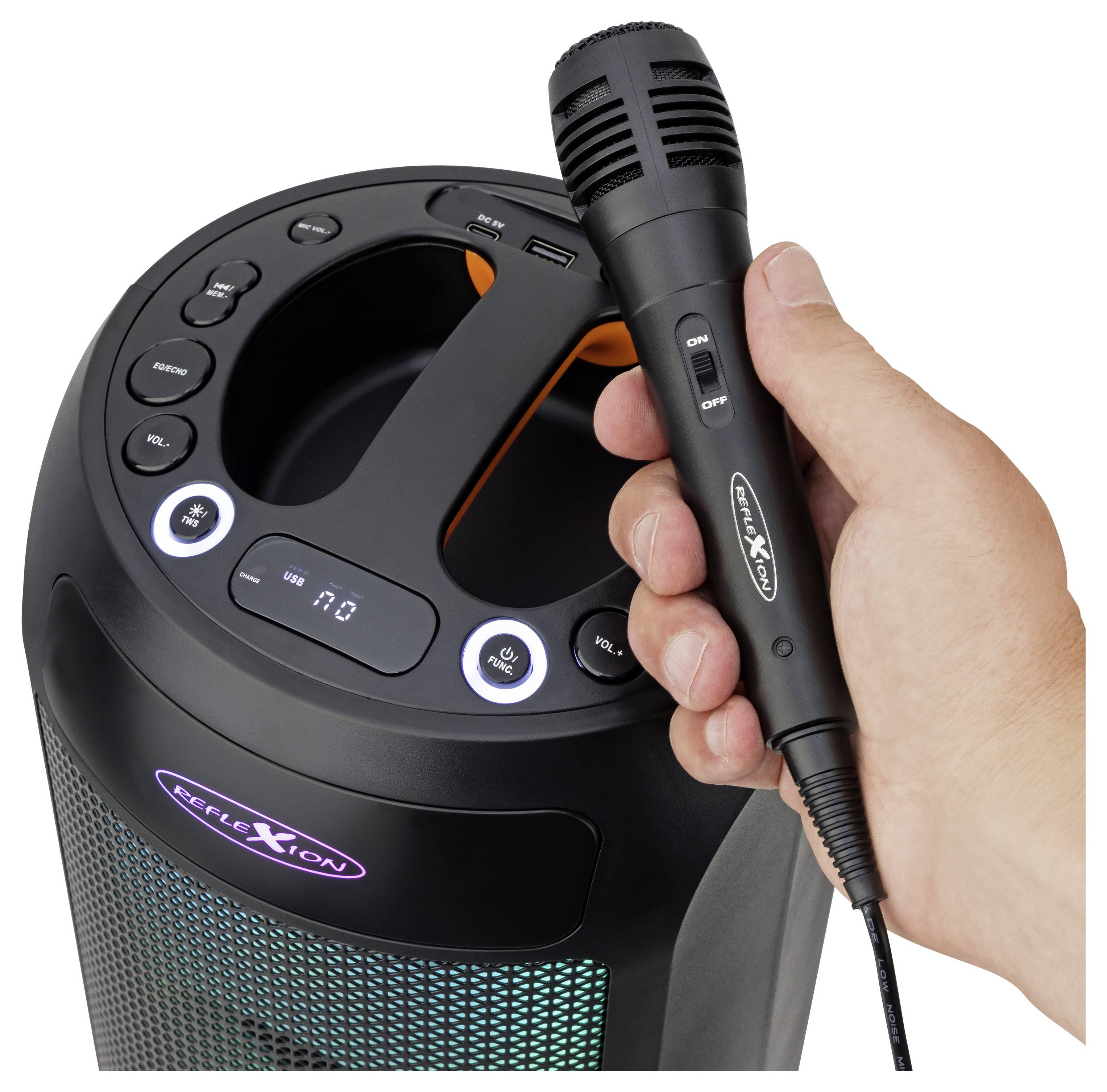 Eine Hand hält ein Mikrofon vor einem Lautsprecher mit Bedienungstasten und einer LED-Anzeige, was einen Karaoke-Aufbau andeutet.