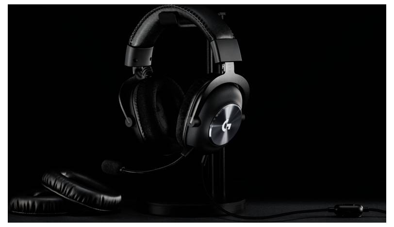 Ein schwarzes Gaming-Headset mit einem metallischen Logo und Mikrofon sitzt auf einem Ständer, mit zusätzlichen Ohrpolstern in der Nähe, vor einem dunklen Hintergrund.
