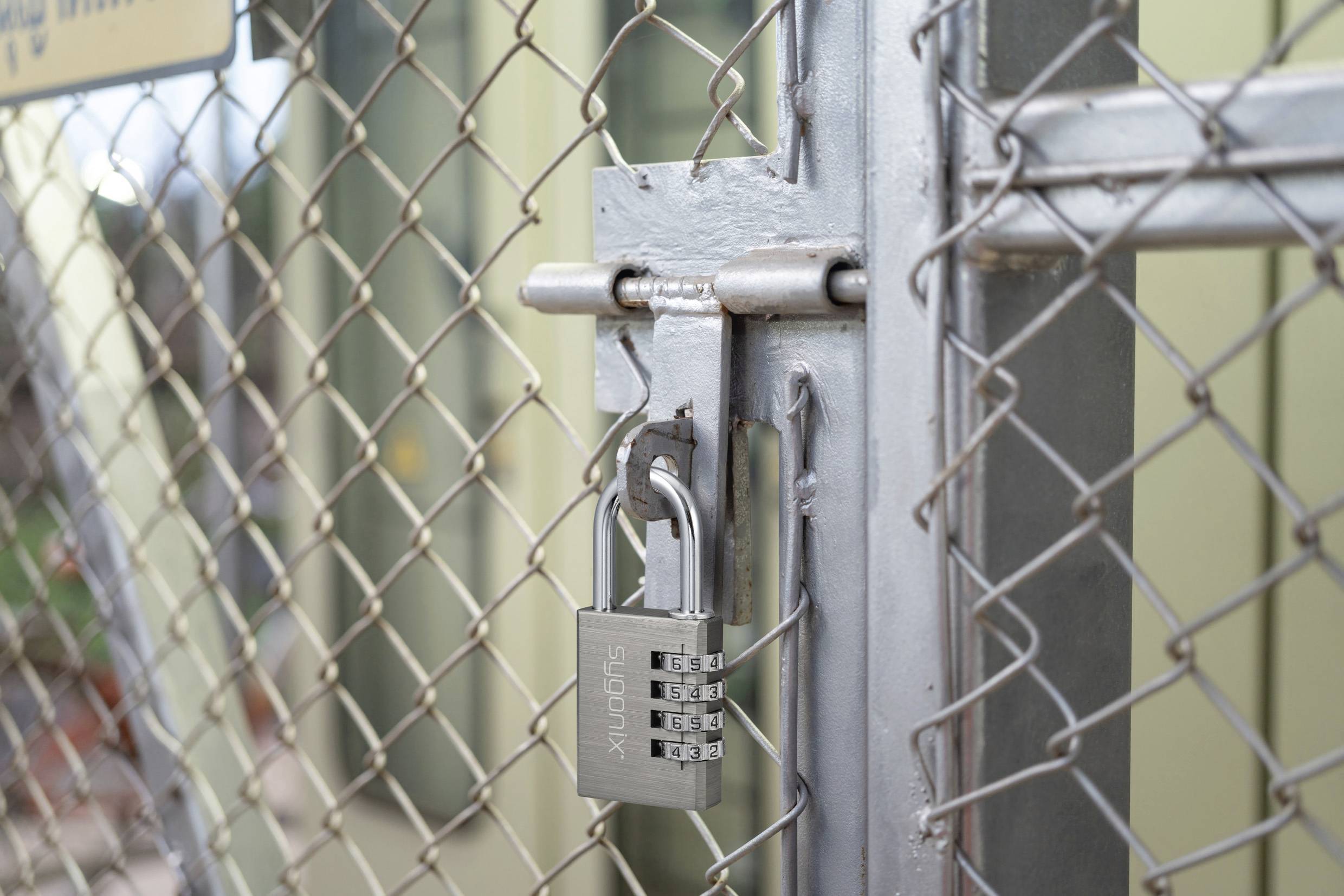 Ein silberner Kombinationsvorhängeschloss sichert ein Maschendrahtor und deutet auf einen eingeschränkten Zugang zu dem eingezäunten Bereich dahinter hin.