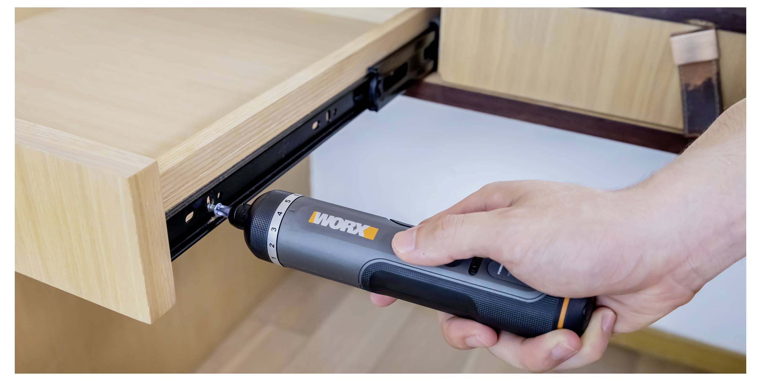 Eine Hand benutzt einen Akku-Schrauber, um eine Schublade an ihren Gleitschienen in einem Holzschrank zu befestigen und damit die Montage von Möbeln in Eigenarbeit zu veranschaulichen.