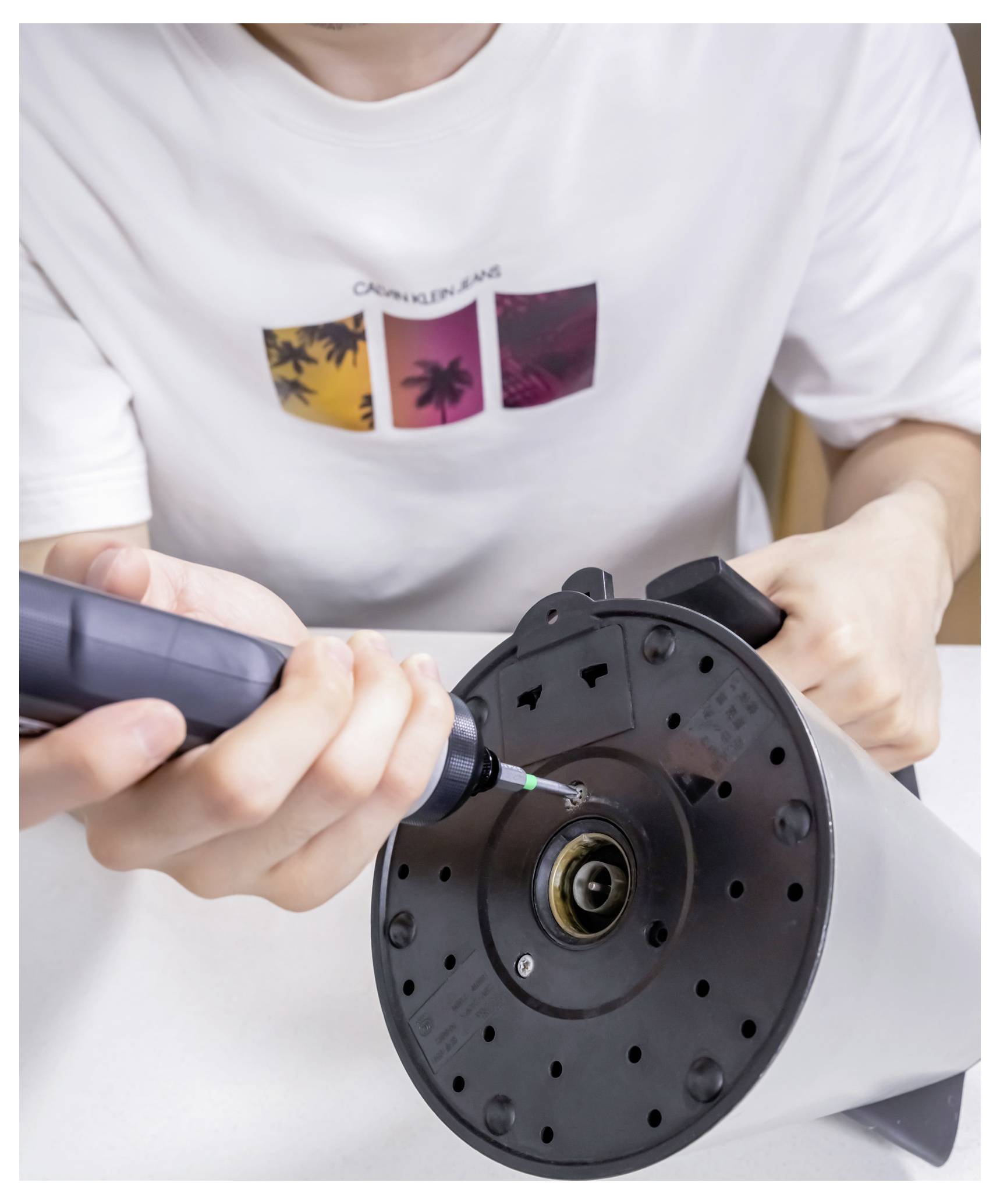 Eine Person in einem weißen T-Shirt benutzt einen Schraubendreher, um ein rundes elektronisches Gerät auf einer weißen Oberfläche zu reparieren oder zusammenzubauen.