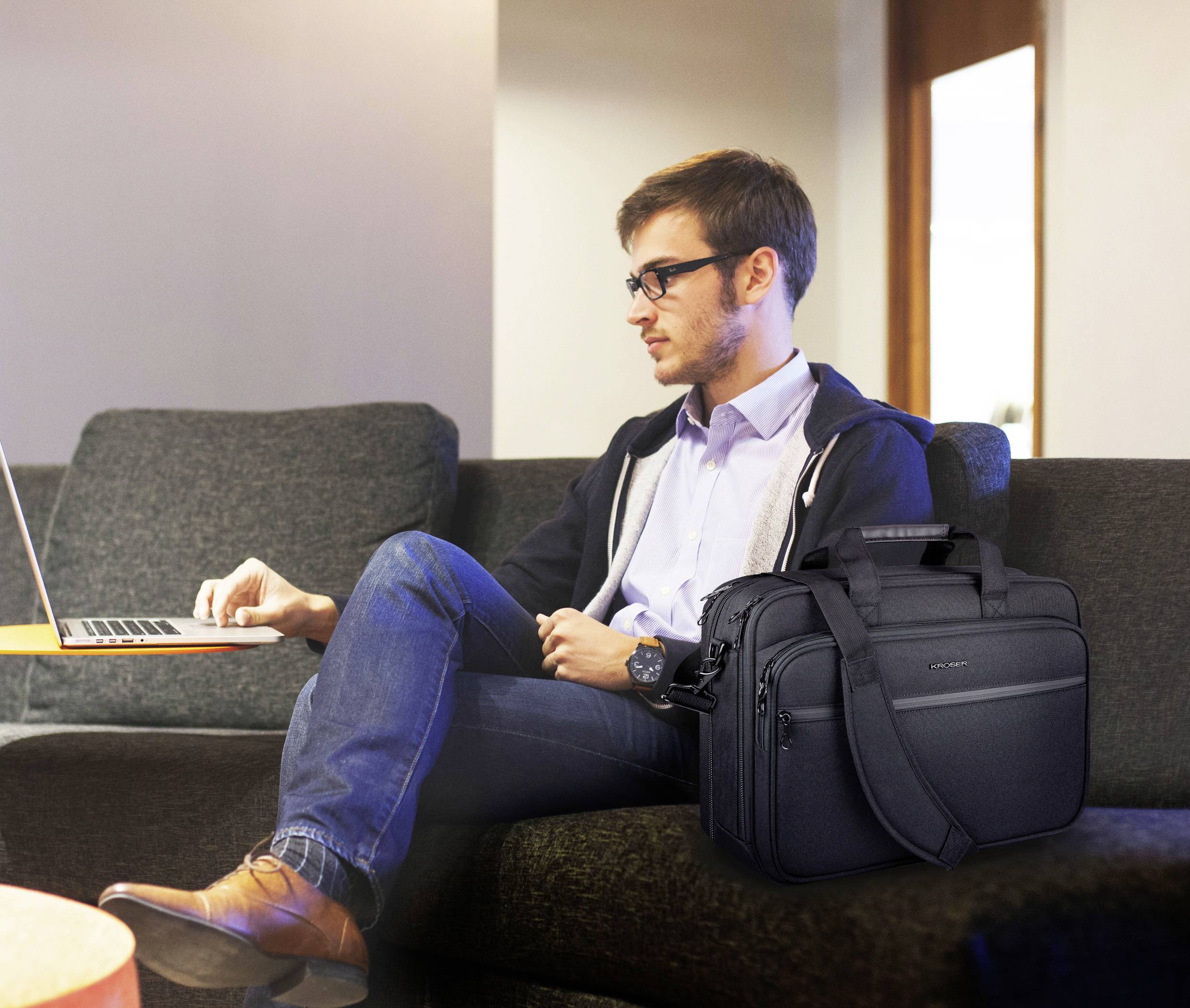 Ein Mann sitzt auf einer Couch und benutzt einen Laptop, neben ihm liegt eine schwarze Laptoptasche.