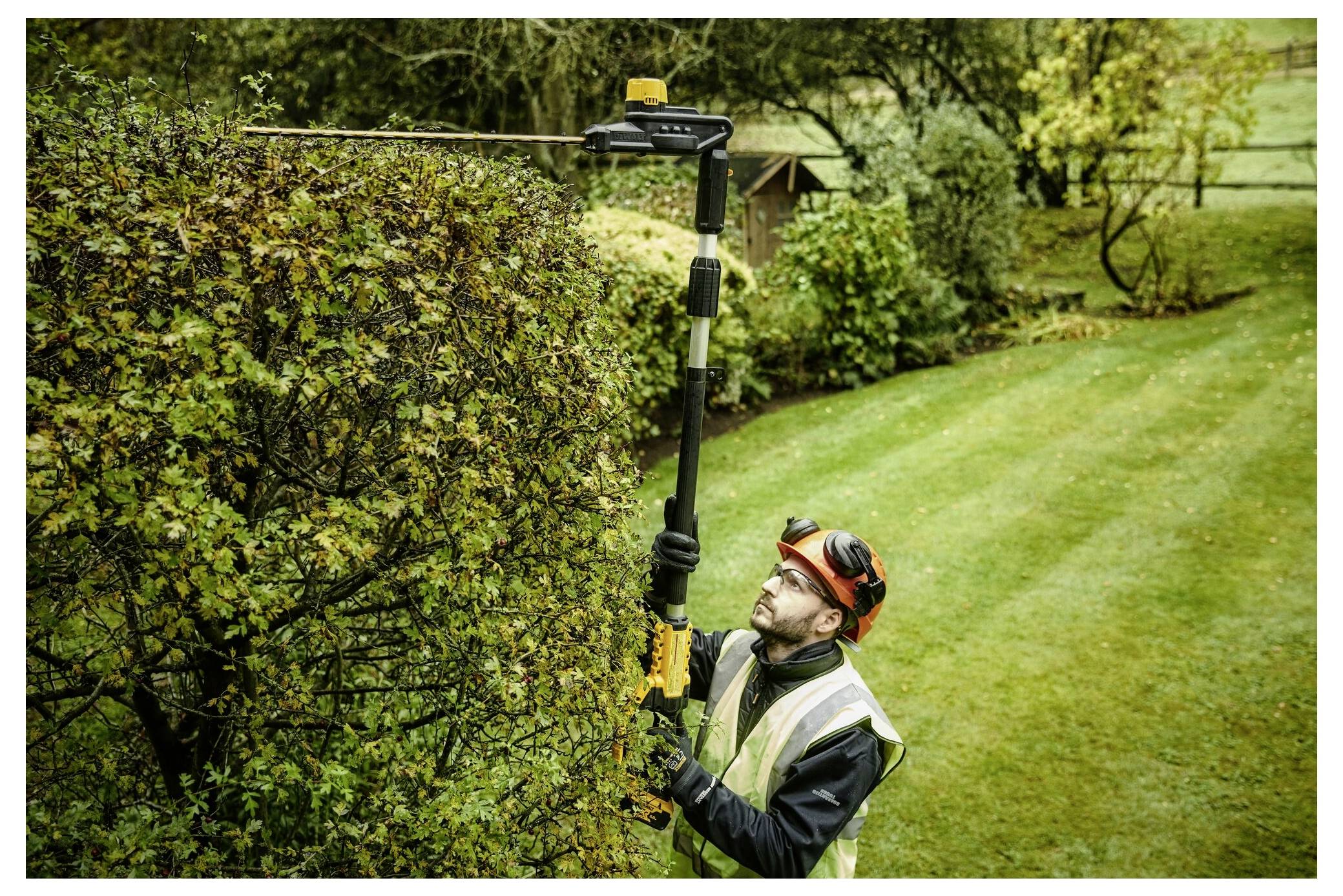 Eine Person in Sicherheitsausrüstung schneidet hohe Hecken in einem Garten mit einem elektrischen Heckenschneider, umgeben von üppigem Grün und Bäumen.