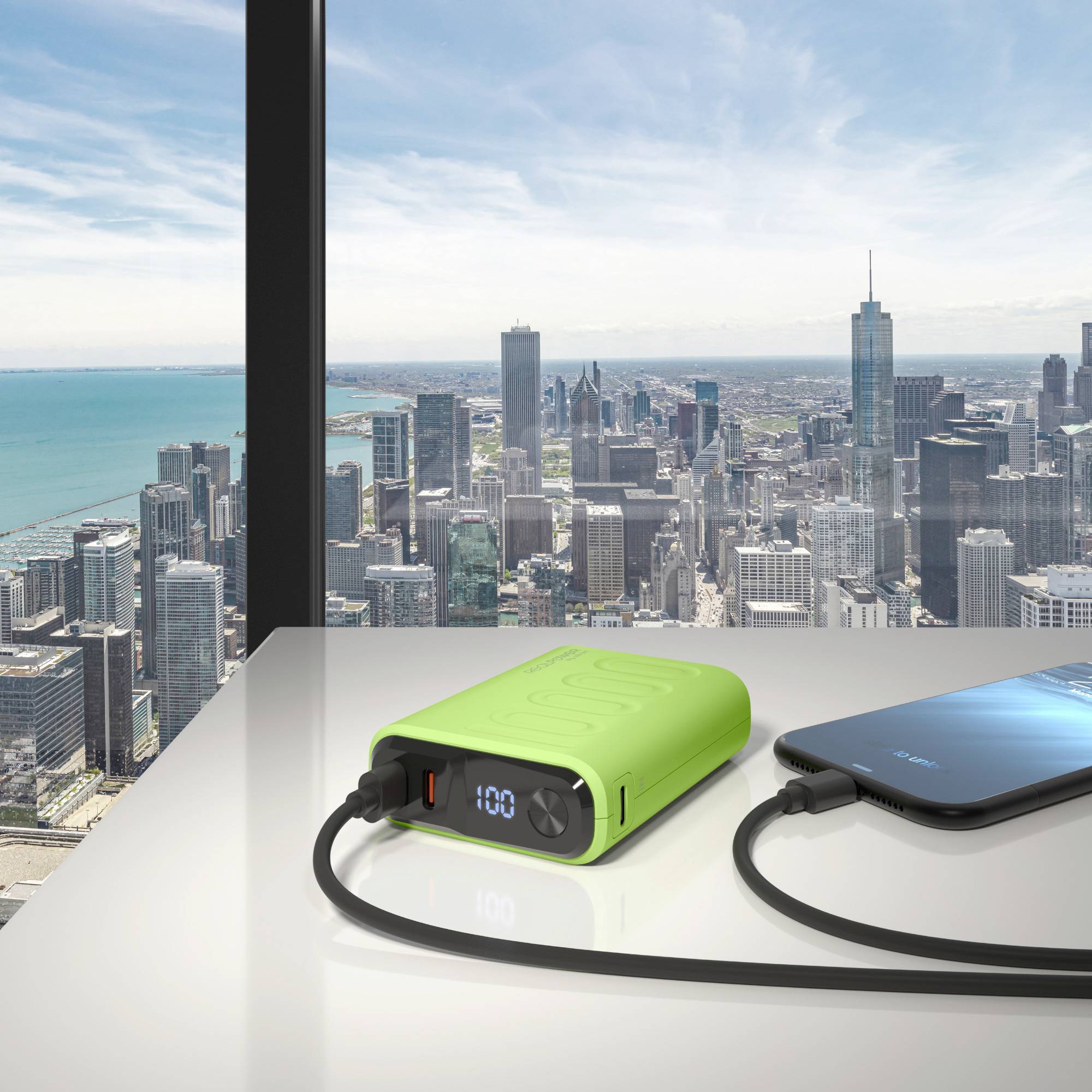 Ein grünes Powerbank, das ein Smartphone auf einem Tisch neben einem Fenster mit Blick auf die Skyline einer Stadt an einem sonnigen Tag lädt.