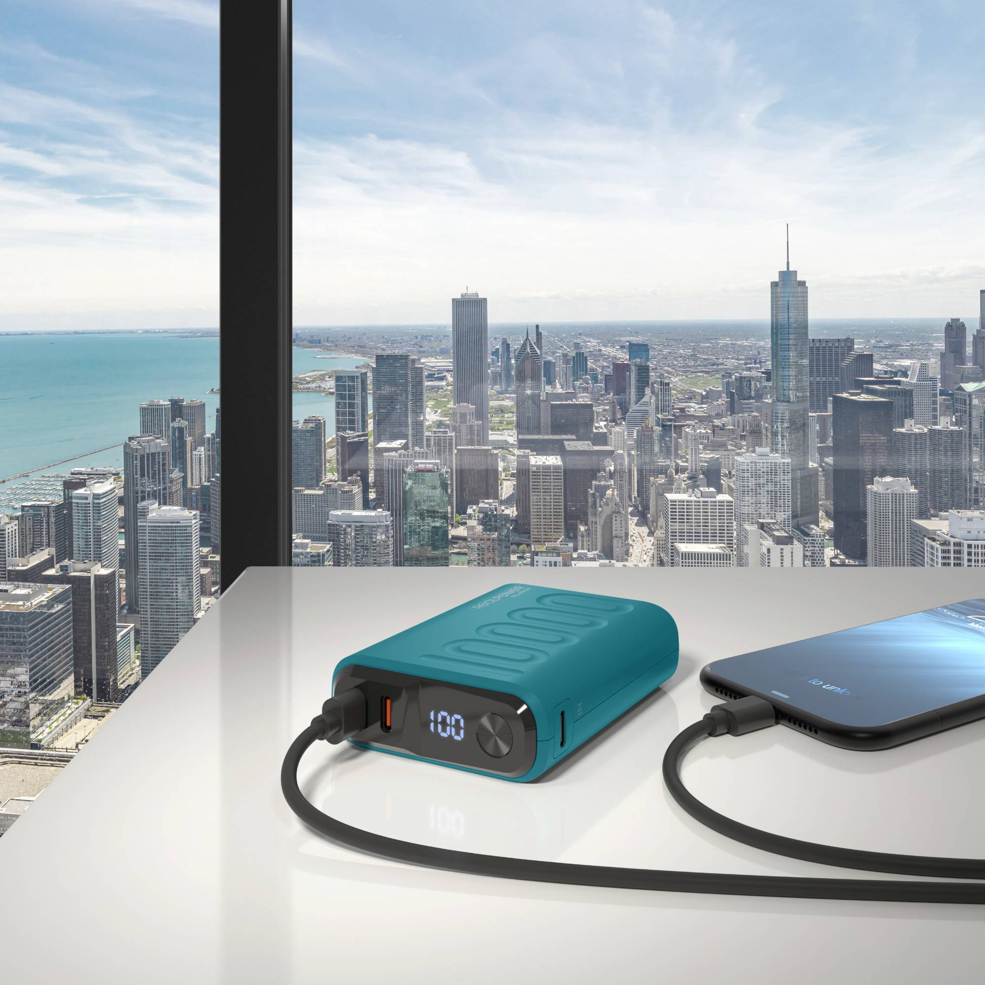 Eine Powerbank lädt ein Smartphone auf einem Tisch mit Blick auf eine Stadtlandschaft im Hintergrund durch große Fenster.