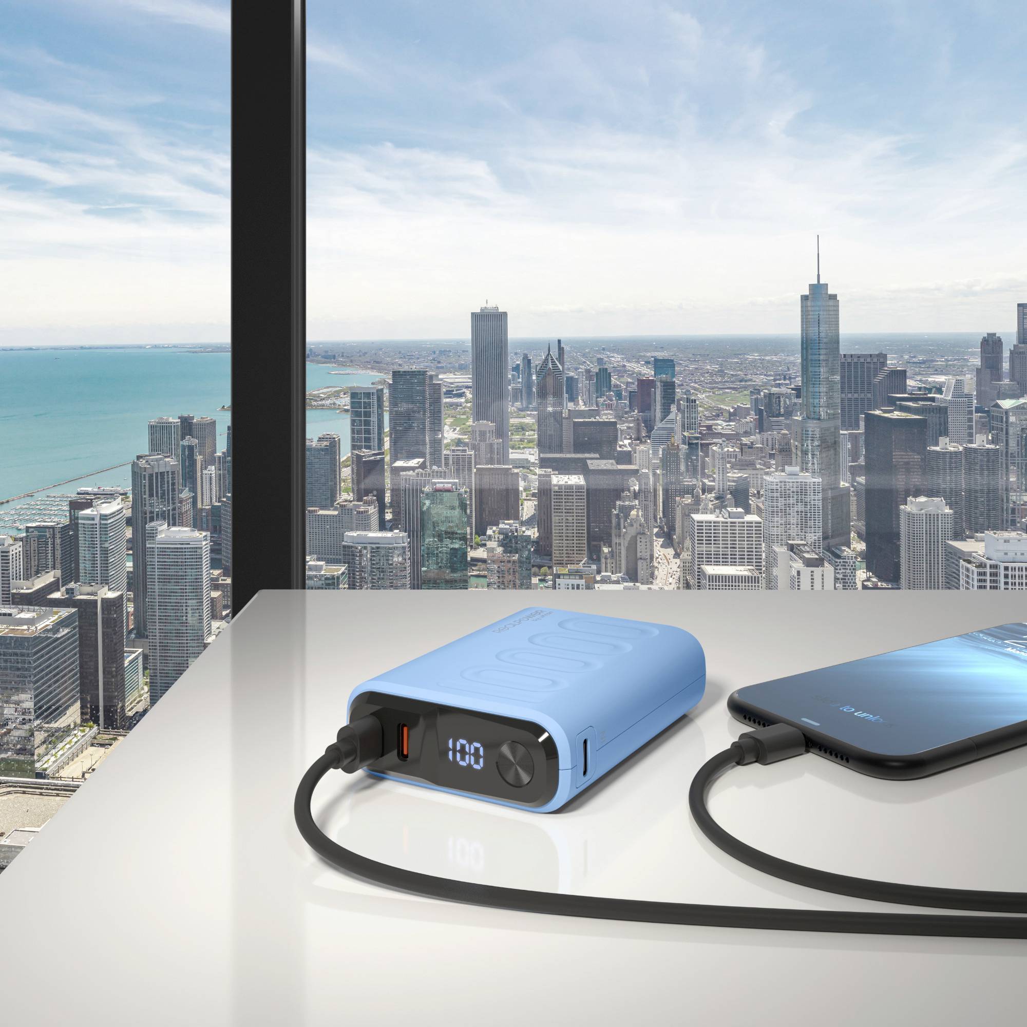 Ein blauer digitaler Powerbank lädt ein Smartphone auf einem Tisch neben einem großen Fenster. Das Fenster bietet Blick auf eine weitläufige Stadtlandschaft.