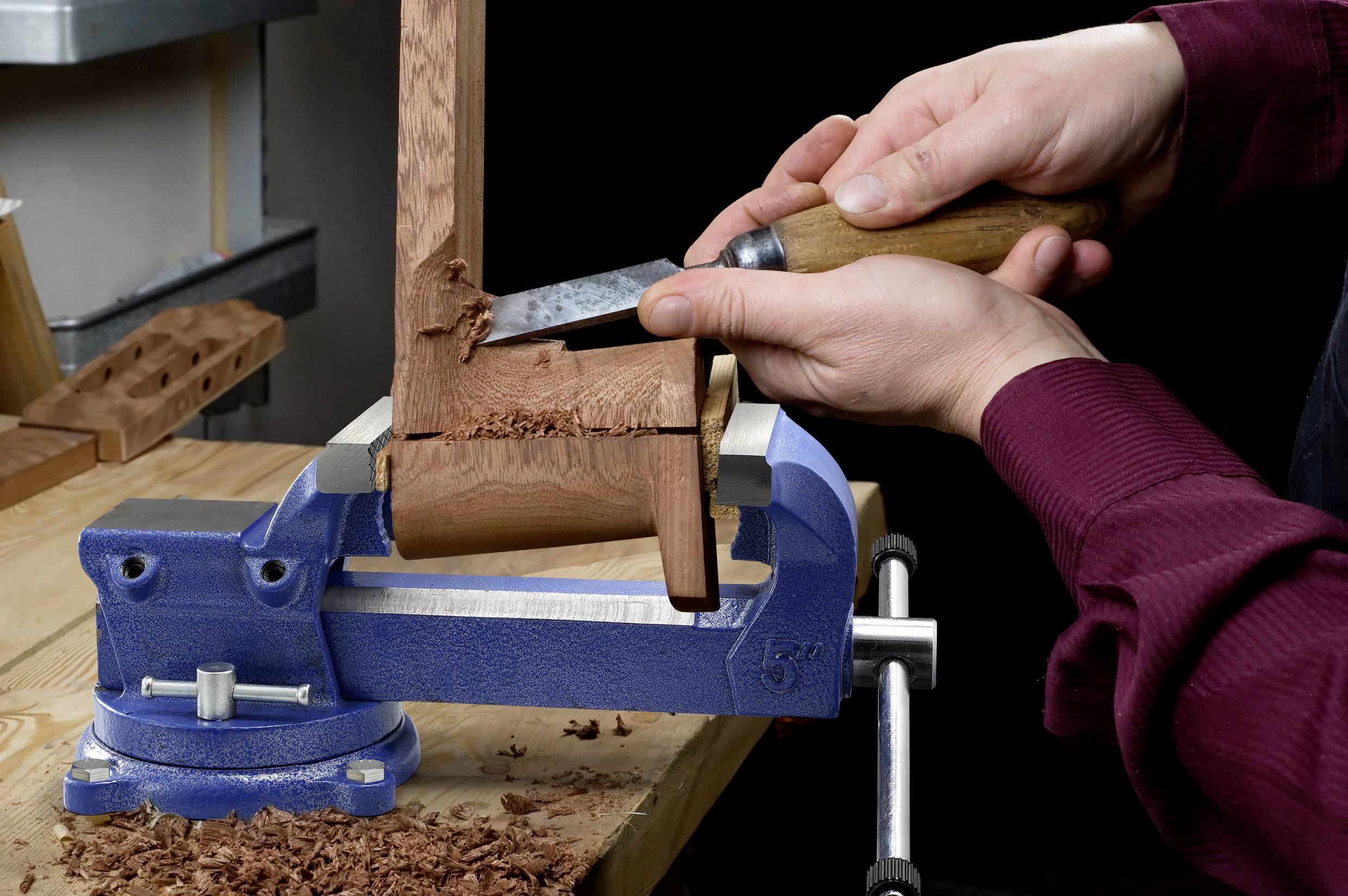 Eine Person verwendet einen Meißel und einen Hammer, um Holz zu bearbeiten, das in einem blauen Schraubstock eingespannt ist und dabei Fertigkeiten in der Holzbearbeitung unter Beweis stellt.