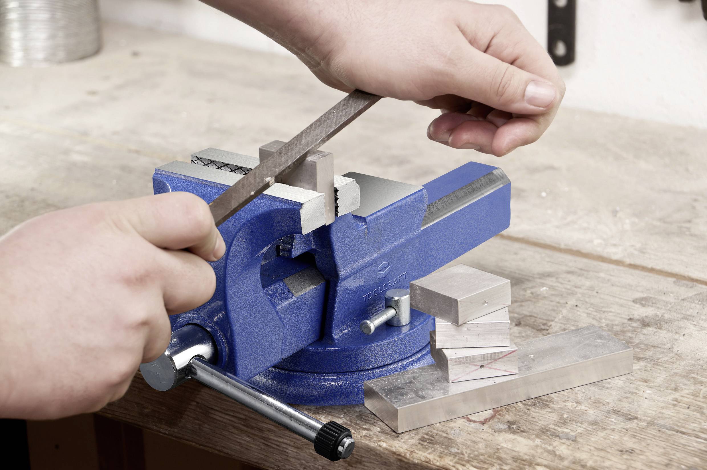 Hände, die eine Metallfeile auf einem Werkstück verwenden, das in einem blauen Schraubstock auf einer Werkbank befestigt ist, was Metallbearbeitung oder Werkzeuggebrauch veranschaulicht.