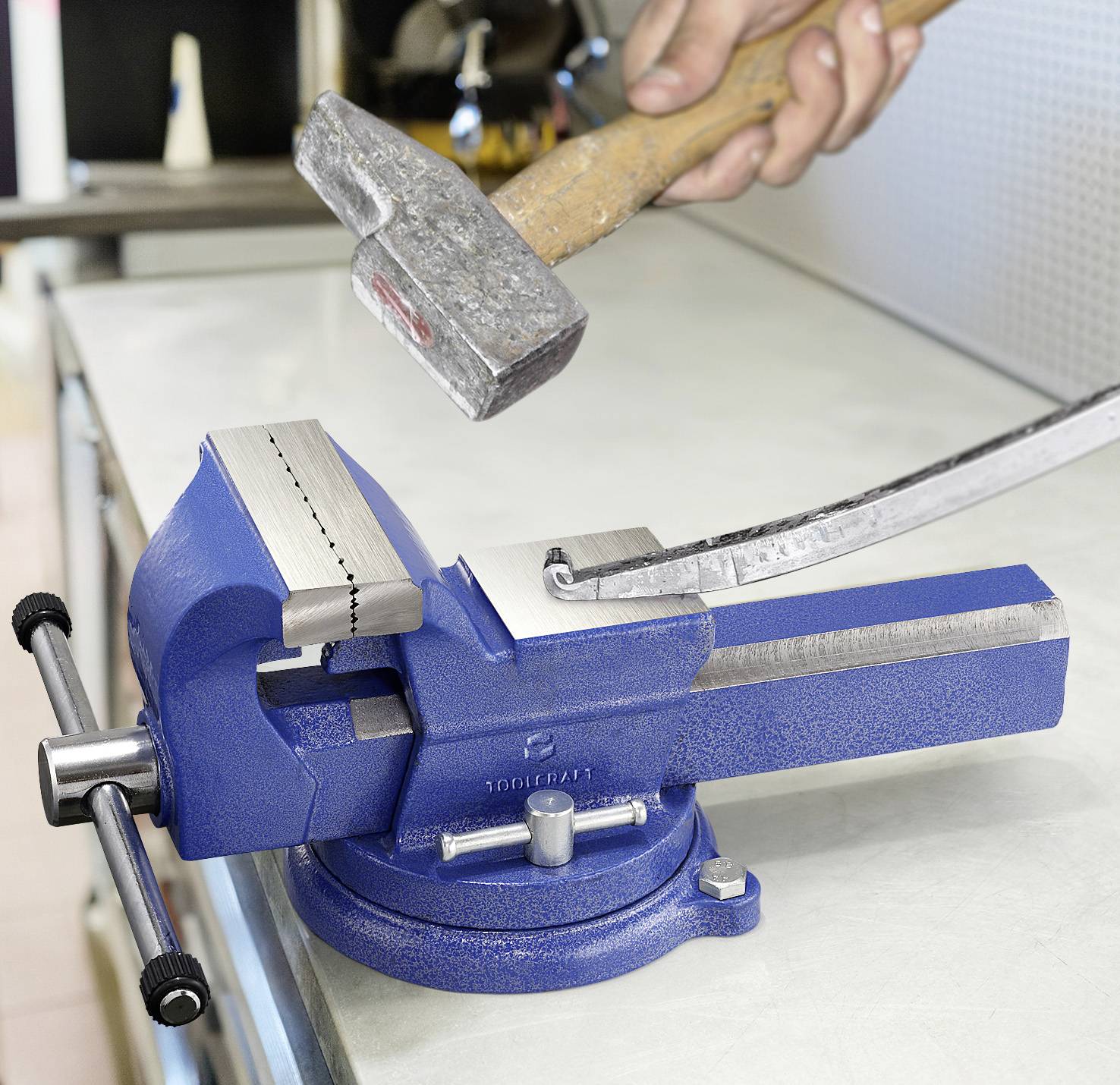Eine blaue Schraubzwinge hält ein metallisches Werkstück auf einer Werkbank, während eine Hand es mit einem Hammer bearbeitet und damit eine Metallbearbeitung andeutet.