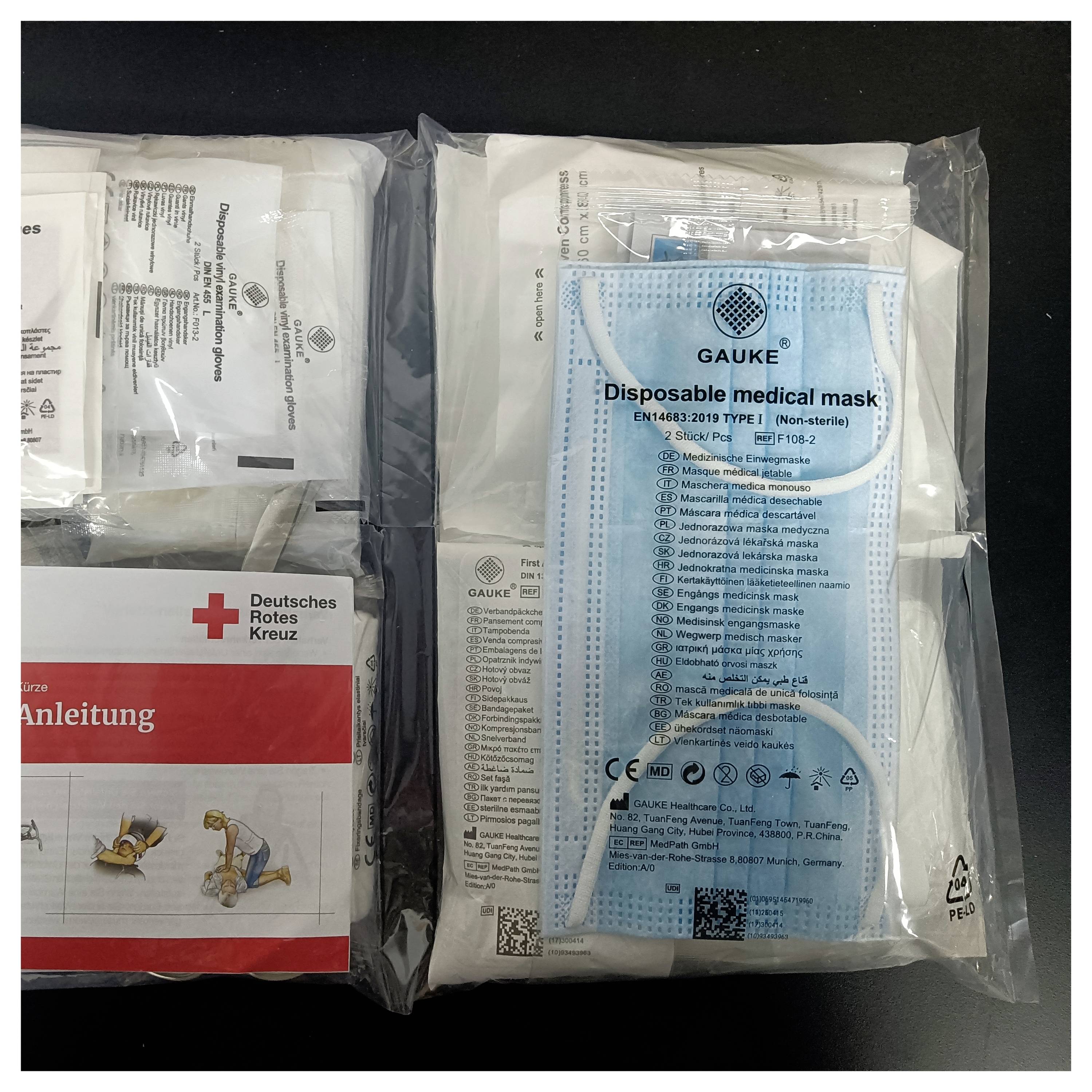 Eine Packung 'Gauke' Einweg-Medizinmasken neben einem Deutschen Roten Kreuz Erste-Hilfe-Set, beide in Plastikverpackungen versiegelt auf einer schwarzen Oberfläche.