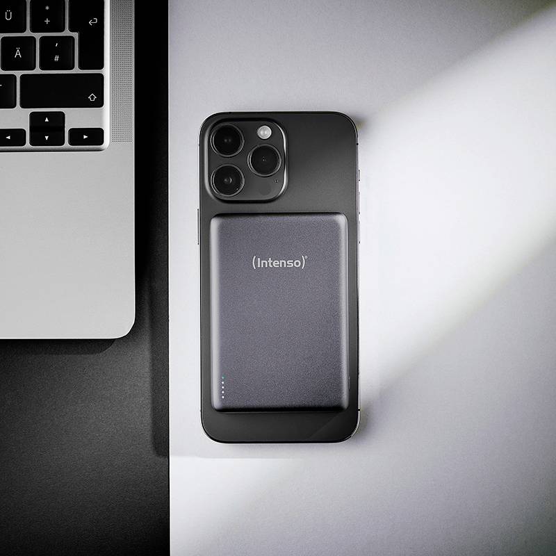 Ein Smartphone auf einem Schreibtisch mit einer rechteckigen, tragbaren Powerbank, die an seiner Rückseite befestigt ist. Ein Teil einer Laptop-Tastatur ist links sichtbar.