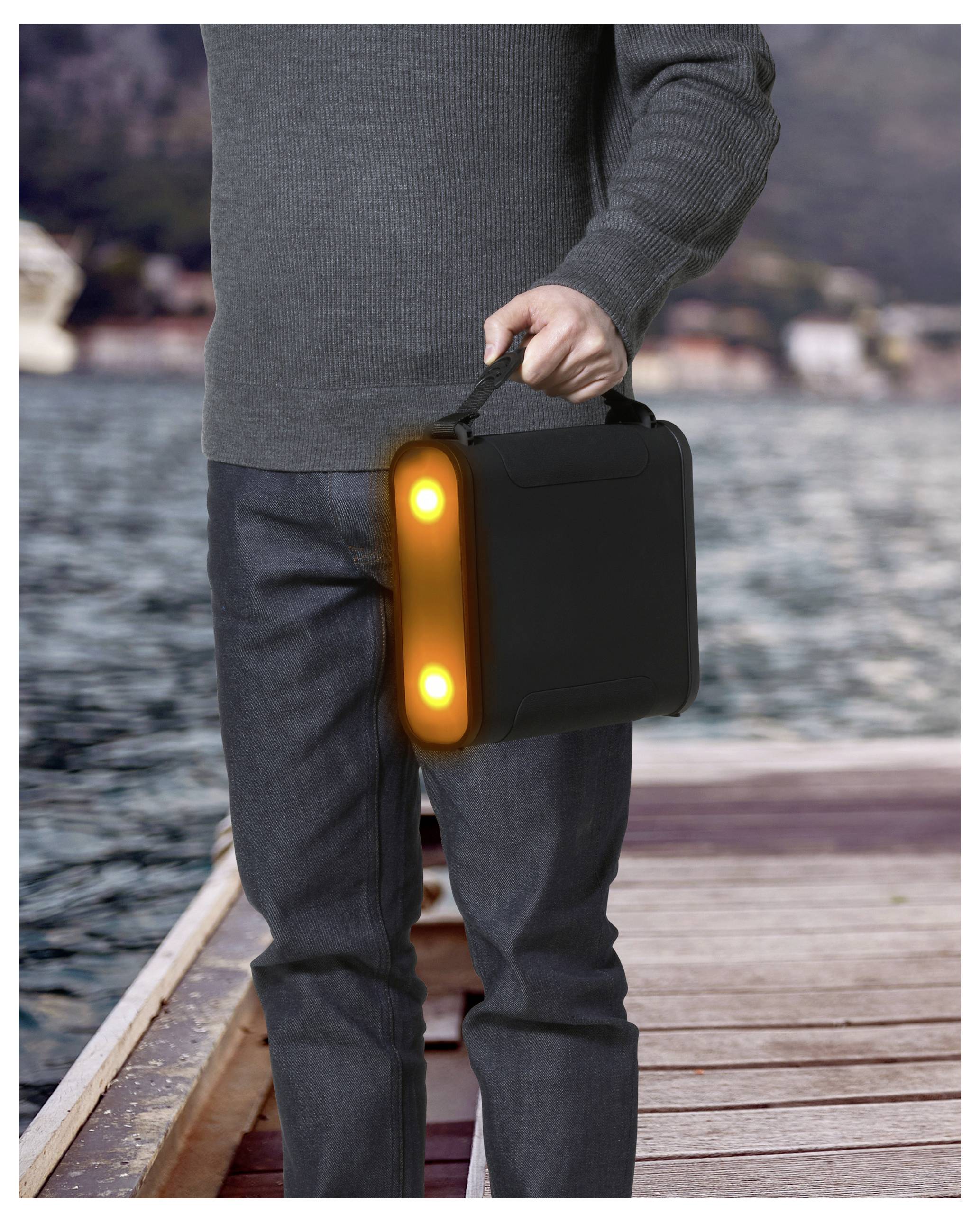 Eine Person geht auf einem Steg und trägt einen schwarzen Aktenkoffer mit zwei leuchtenden orangefarbenen Lichtern vor einem unscharfen Wasser- und Berghintergrund.