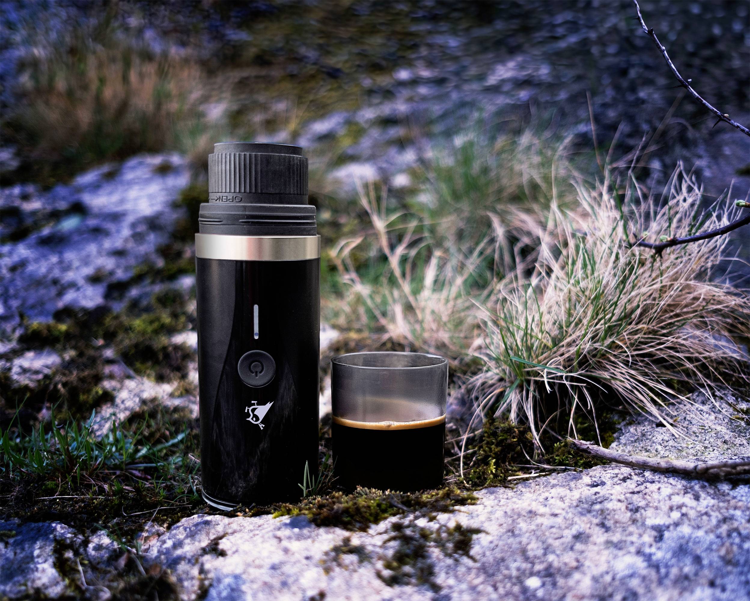 Ein schwarzer Thermobecher und eine halb gefüllte Tasse Kaffee stehen auf einem moosbedeckten Felsen, umgeben von Gras und felsigem Gelände.