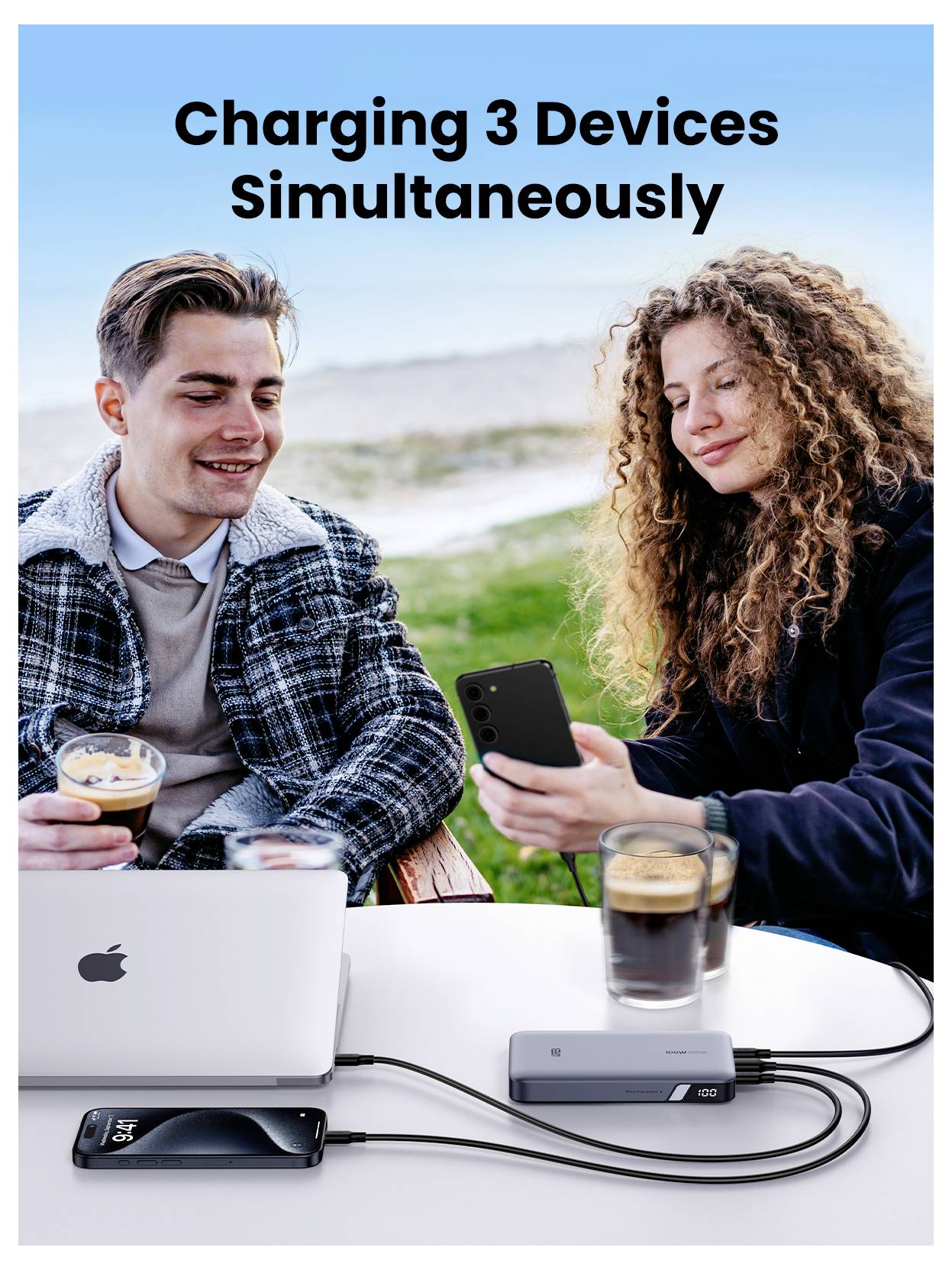 Zwei Personen sitzen im Freien und nutzen einen Laptop und Smartphones. Ein Ladehub versorgt drei Geräte mit Strom und veranschaulicht das gleichzeitige Laden.