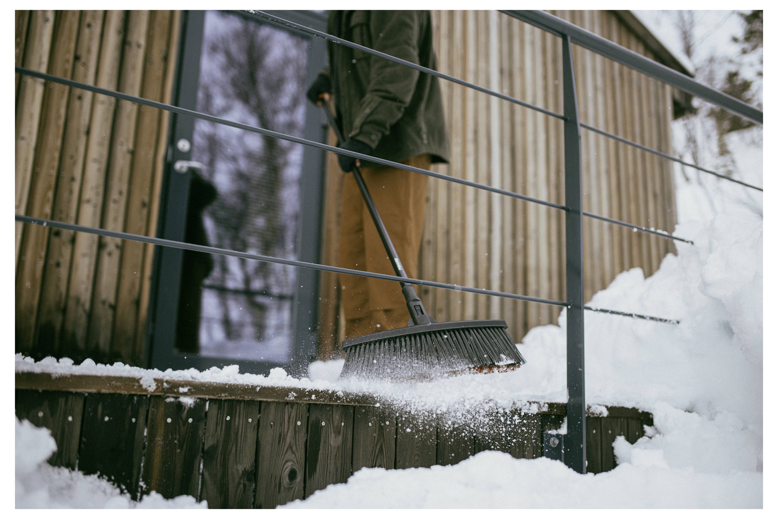 Eine Person, die mit einem Besen Schnee von den Holztreppenstufen einer Hütte fegt, gekleidet in eine Jacke und Hose. Schnee umgibt das Gebiet, Bäume im Hintergrund.