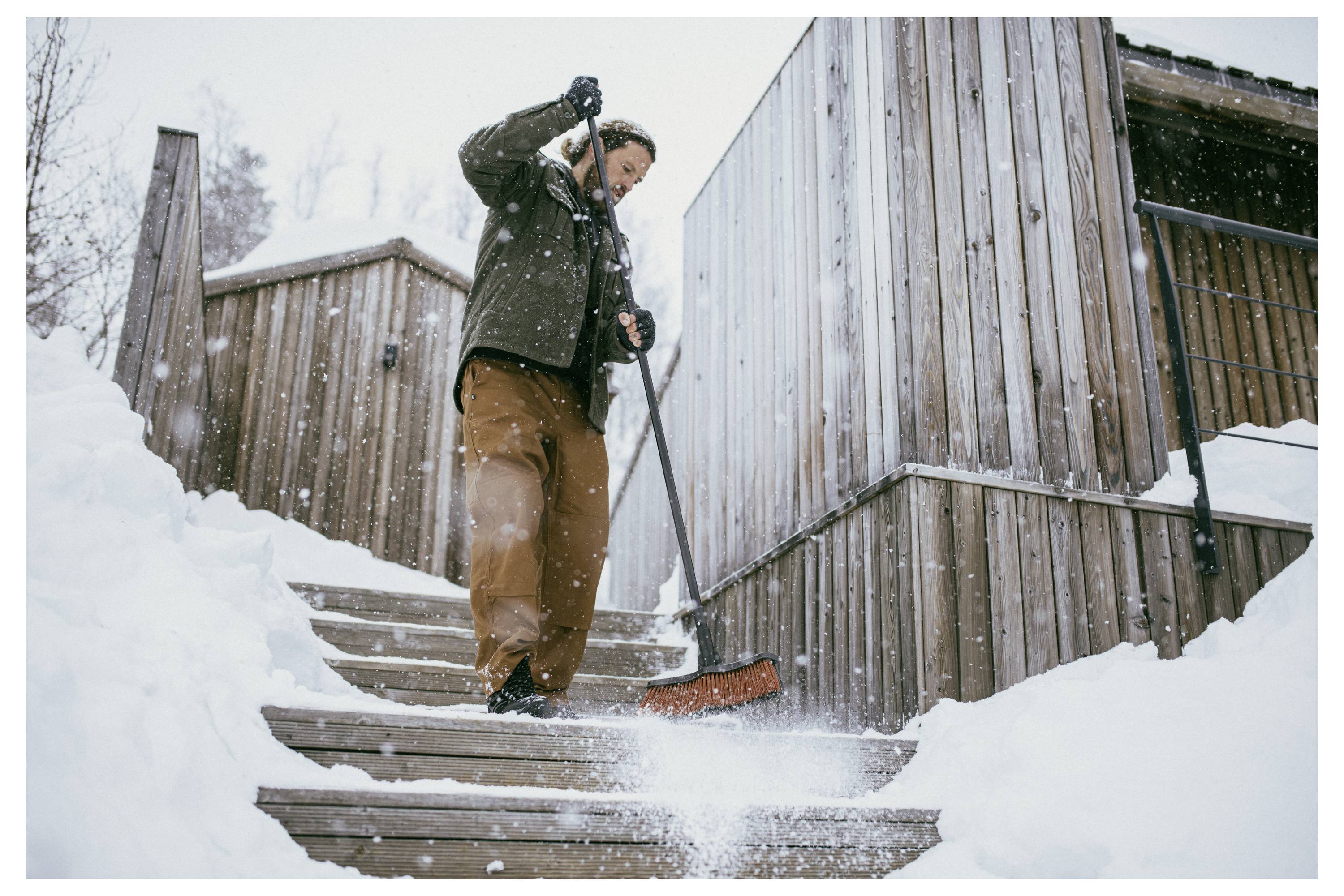 Eine Person schaufelt Schnee von hölzernen Stufen neben einer rustikalen Hütte im Winter, umgeben von fallendem Schnee, und schafft dabei einen sicheren Weg.
