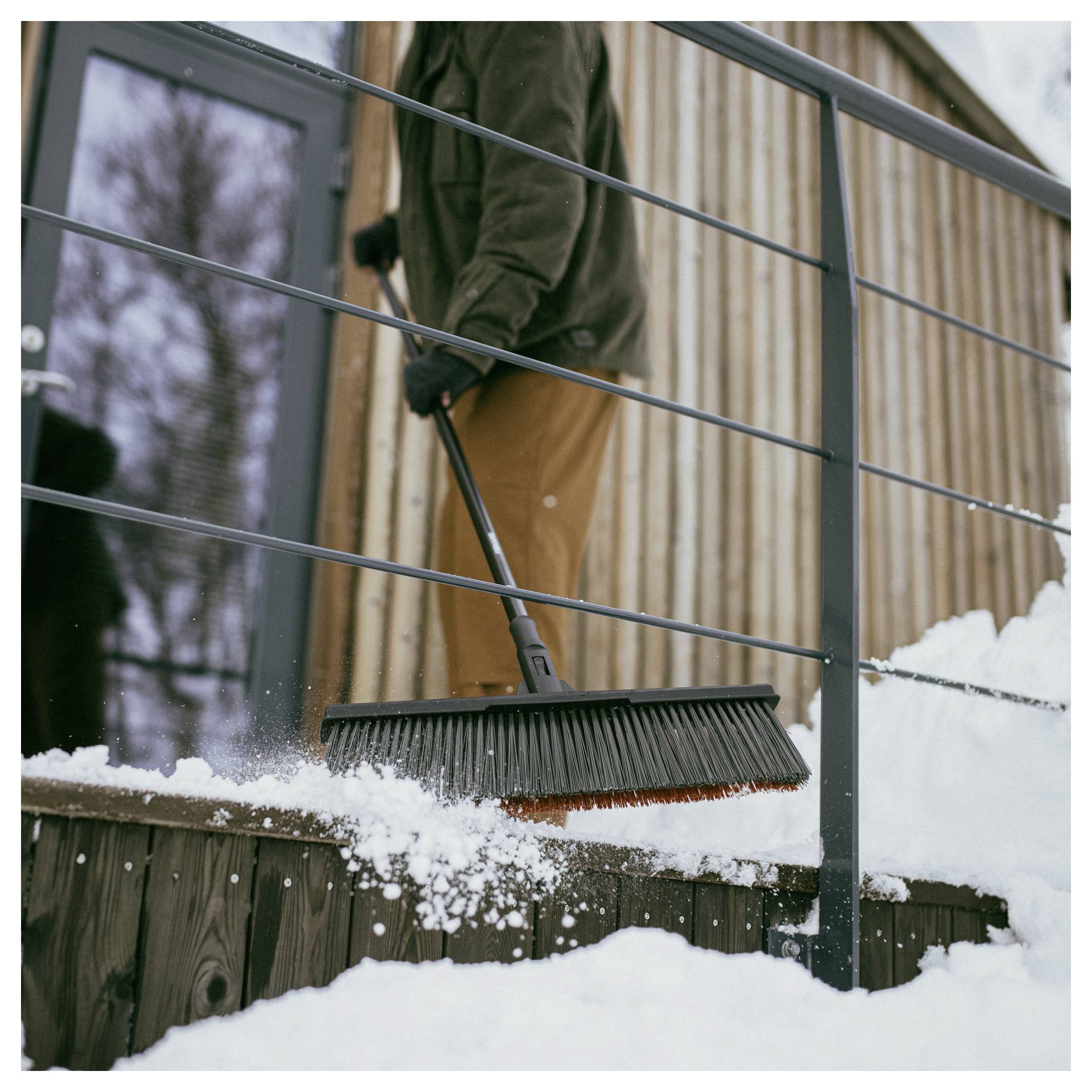 Eine Person in Winterkleidung kehrt mit einem breiten Besen Schnee von hölzernen Stufen neben einem rustikalen Holzgebäude mit Metallgeländern.