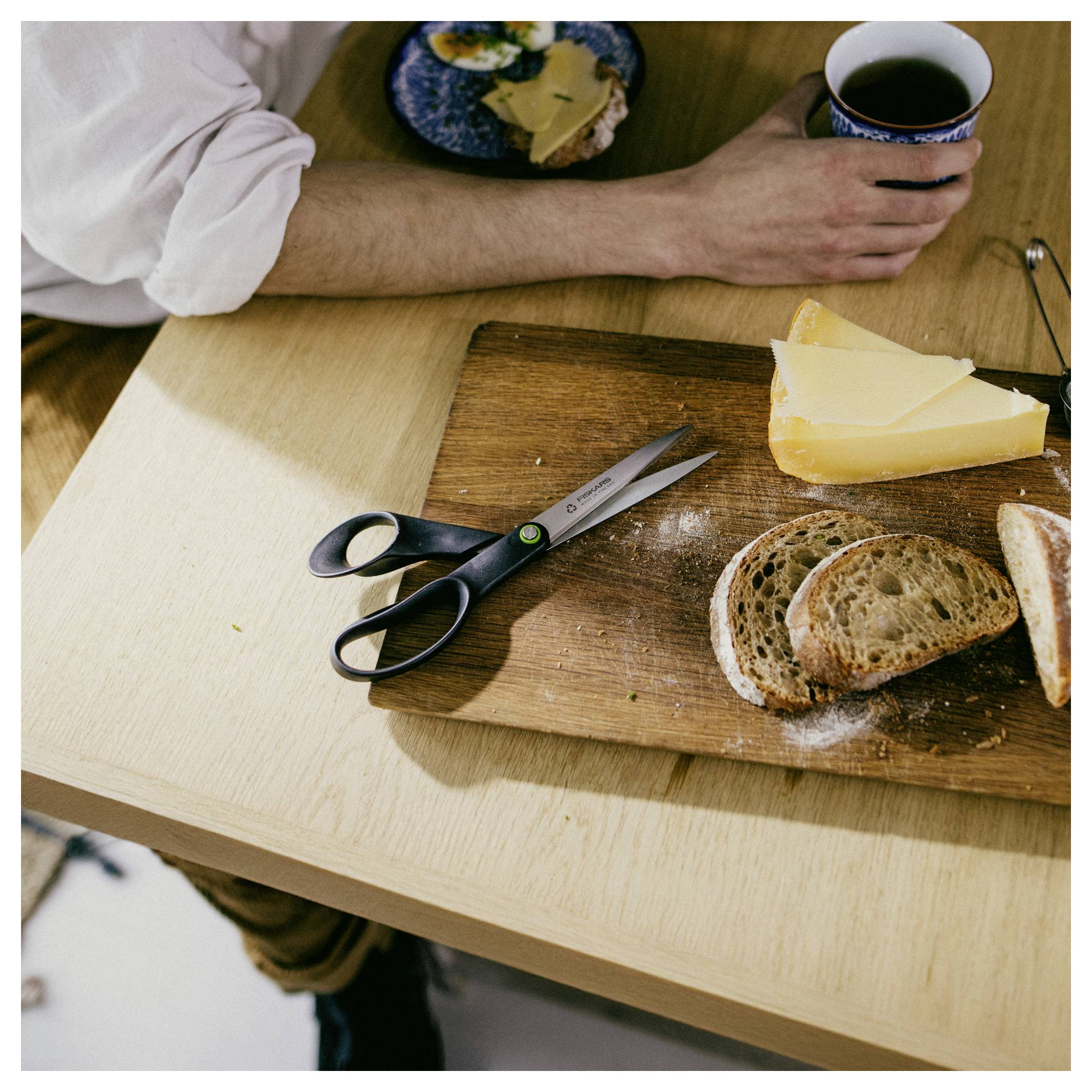 Eine Person sitzt an einem Tisch mit einer Tasse Kaffee, Brot, Käse auf einem Schneidebrett und einer Schere.