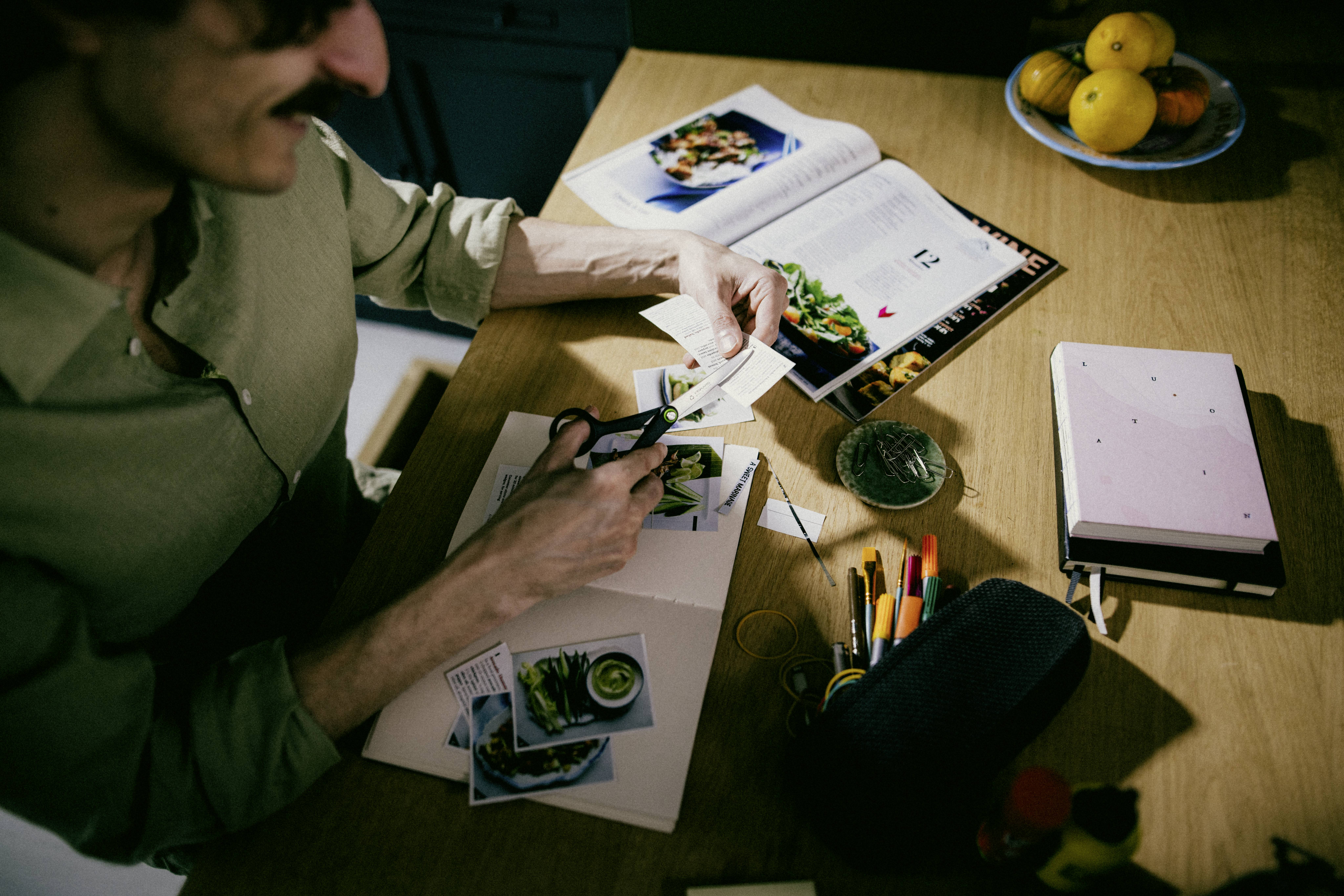 Eine Person schneidet gedruckte Fotos an einem Tisch mit einem aufgeschlagenen Kochbuch, Bleistiften und einer Obstschale, was auf ein kreatives oder kulinarisches Projekt hindeutet.