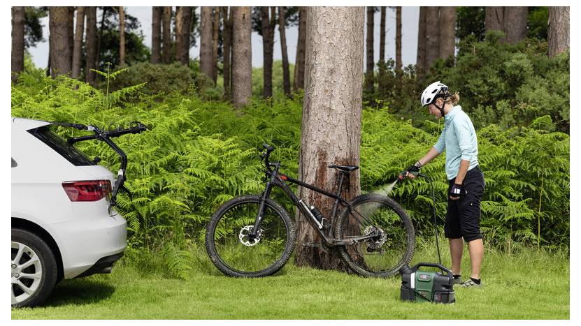 Eine Person in Fahrradbekleidung reinigt ein schwarzes Fahrrad, das an einen Baum gelehnt ist, neben einem weißen Auto. Der Hintergrund ist von üppiger Vegetation und hohen Bäumen geprägt.