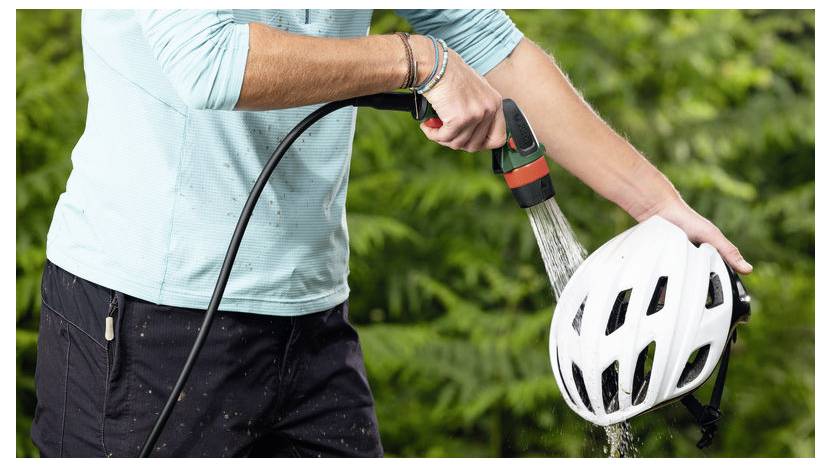 Eine Person benutzt einen Schlauch, um einen weißen Fahrradhelm zu waschen, mit grünem Laub im Hintergrund.