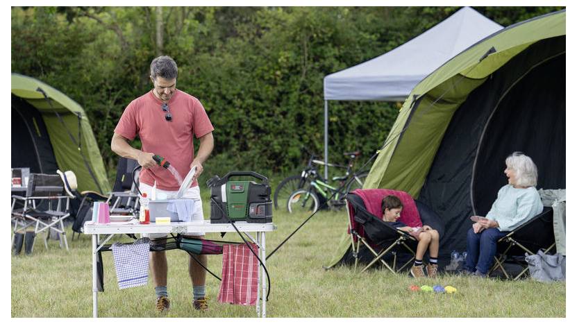Ein Mann bereitet Essen an einem Campingtisch vor, während eine Frau und ein Kind in der Nähe auf Stühlen außerhalb der Zelte sitzen, umgeben von Campingausrüstung.