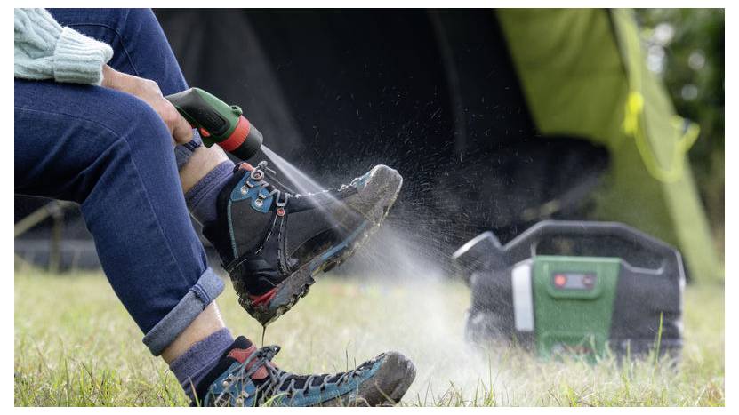 Eine Person, die schlammige Wanderstiefel mit einer tragbaren Wassersprühflasche reinigt, neben einem grünen Zelt auf Gras. Eine mobile Stromversorgungseinheit befindet sich in der Nähe.