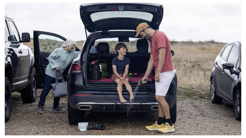 Ein Mann wäscht die Füße eines Kindes mit einem Gartenschlauch am offenen Kofferraum eines Autos. Eine ältere Person steht in der Nähe, hält eine Tasche und beobachtet die Szene.