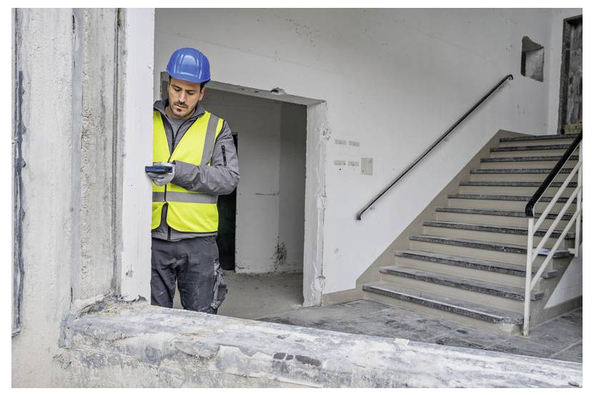 Ein Bauarbeiter in einer Sicherheitsweste und mit Schutzhelm untersucht ein Gerät in einem teilweise renovierten Gebäude neben einer Treppe.