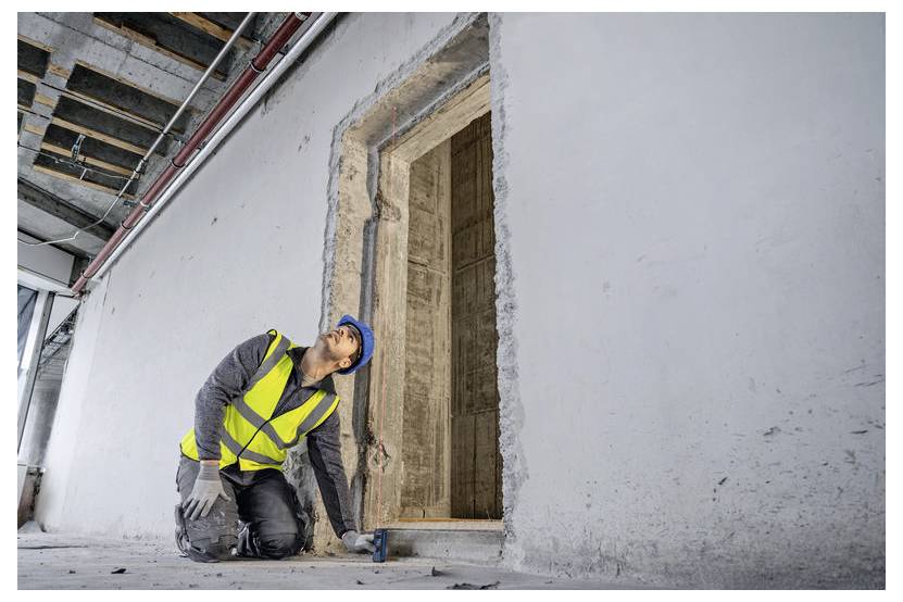 Ein Bauarbeiter in einer gelben Warnweste und einem blauen Schutzhelm untersucht einen Türrahmen in einem unfertig Gebäude und prüft dessen Ausrichtung und Stabilität.