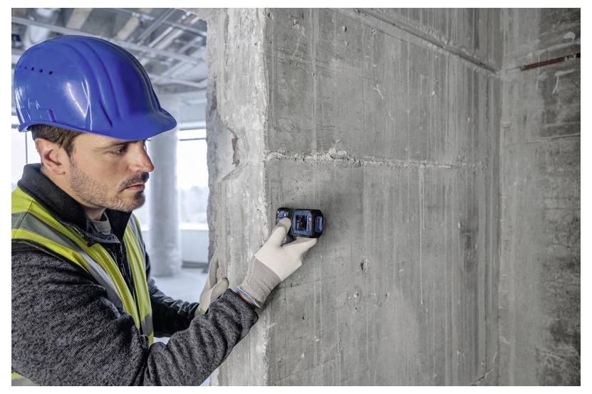Ein Bauarbeiter mit blauem Schutzhelm und Sicherheitsweste verwendet ein Messwerkzeug an einer Betonwand in einem sich im Bau befindlichen Gebäude.