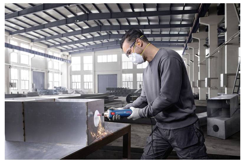 Eine Person mit Sicherheitsausrüstung benutzt ein Elektrowerkzeug, um Metall in einer Industriewerkstatt zu schneiden, wobei Funken aus dem Schneidbereich sprühen.