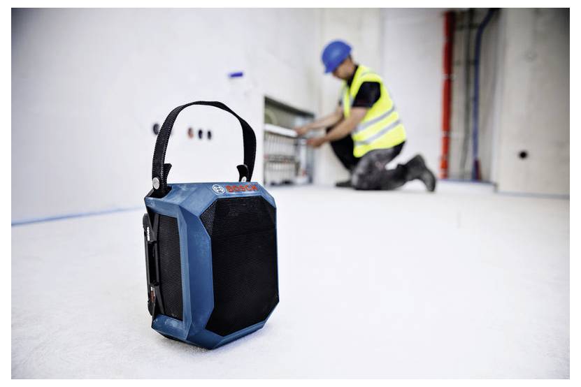 Ein tragbarer blauer Lautsprecher steht im Vordergrund im Fokus, während ein Bauarbeiter in Warnweste und Helm im Hintergrund unscharf ist.