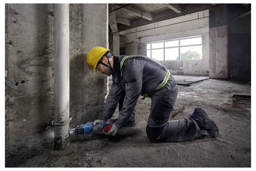 Ein Bauarbeiter in Schutzausrüstung, einschließlich Schutzhelm, kniet und benutzt ein Elektrowerkzeug, um an einem Rohr in einem unfertig gebauten Gebäude zu arbeiten.