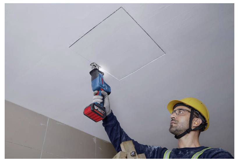 Ein Bauarbeiter mit einem gelben Helm benutzt ein Elektrowerkzeug, um ein Quadrat in einer Deckenplatte auszuschneiden. Er blickt nach oben und ist konzentriert bei der Arbeit.