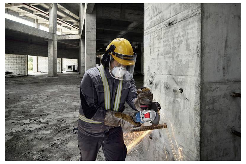 Ein Bauarbeiter schleift eine Betonwand in einer teilweise fertiggestellten Struktur und trägt Schutzausrüstung, einschließlich Helm, Schutzbrille und Handschuhen.