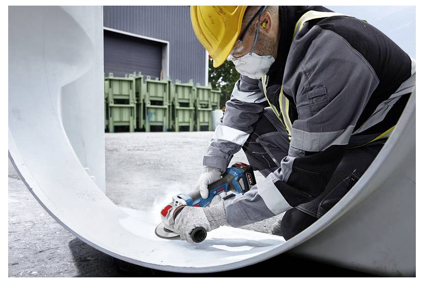 Ein Arbeiter in Sicherheitsausrüstung bedient ein Elektrowerkzeug an einem großen Rohr in einer industriellen Umgebung, mit grünen Behältern im Hintergrund.