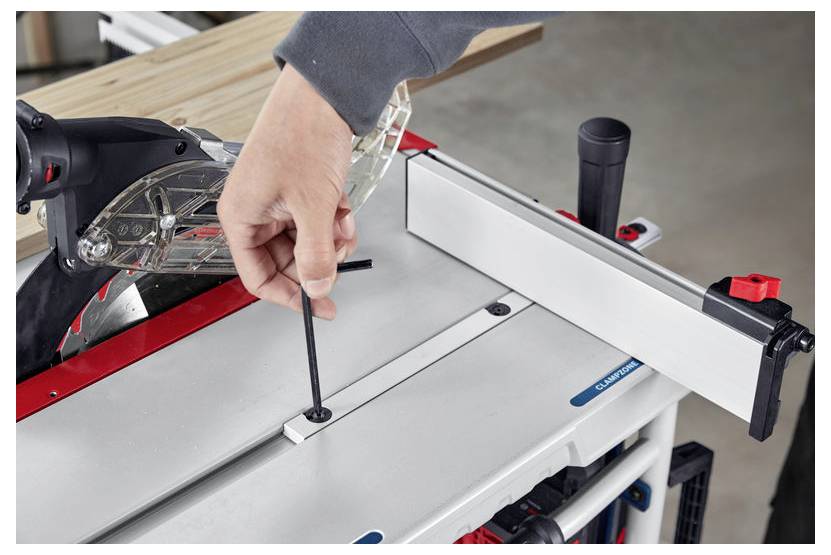 Eine Person justiert ein Tischkreissägeblatt mit einem Sechskantschlüssel und konzentriert sich dabei auf Wartung oder Einrichtung. Die Tischoberfläche ist metallfarbig und verfügt über eine Schutzvorrichtung.