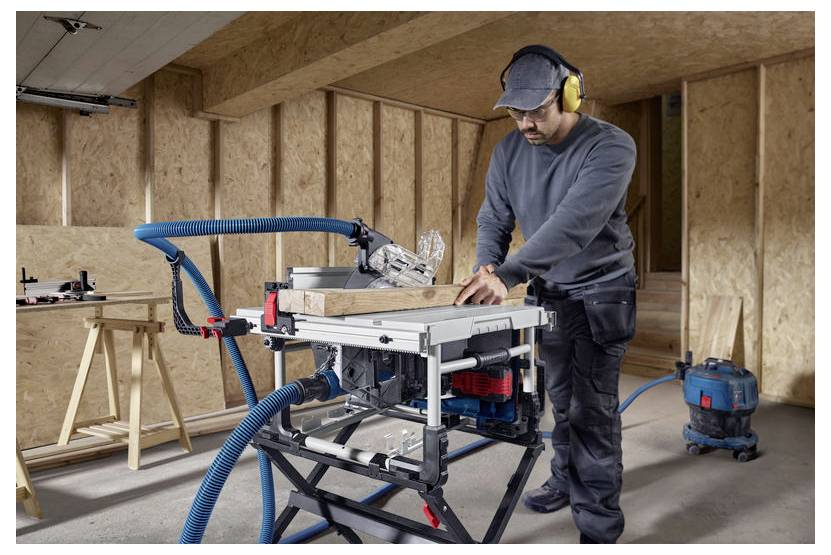 Mann, der in einer Holzwerkstatt eine Kreissäge benutzt und Sicherheitsausrüstung wie Handschuhe und Gehörschutz trägt. Ein Staubsaugerschlauch ist zur Staubkontrolle angeschlossen.