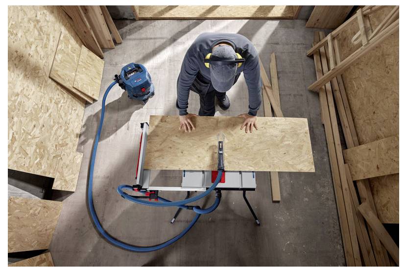 Eine Person mit Mütze und Ohrenschützern sägt ein Holzbrett auf einer Werkbank mit angeschlossenem Staubsauger, umgeben von Holzplatten.