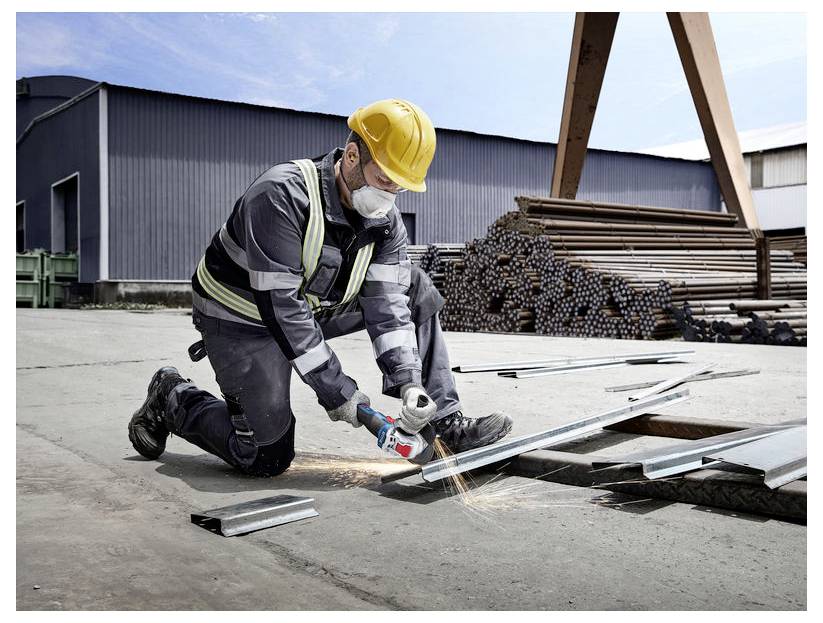 Ein Bauarbeiter mit Sicherheitsausrüstung kniet und verwendet eine Winkelschleiferin, um Metallbleche zu schneiden, während im Hintergrund Stapel von Metallstangen zu sehen sind.