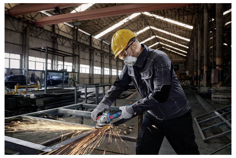 Ein Arbeiter in Sicherheitsausrüstung benutzt einen Winkelschleifer an Metall, wobei Funken entstehen, in einem großen Industrielager mit hohen Decken.