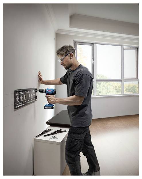 Eine Person mit Sicherheitsbrille verwendet eine Bohrmaschine, um einen schwarzen Rahmen an einer weißen Wand neben einem Fenster in einem hellen Raum zu befestigen.