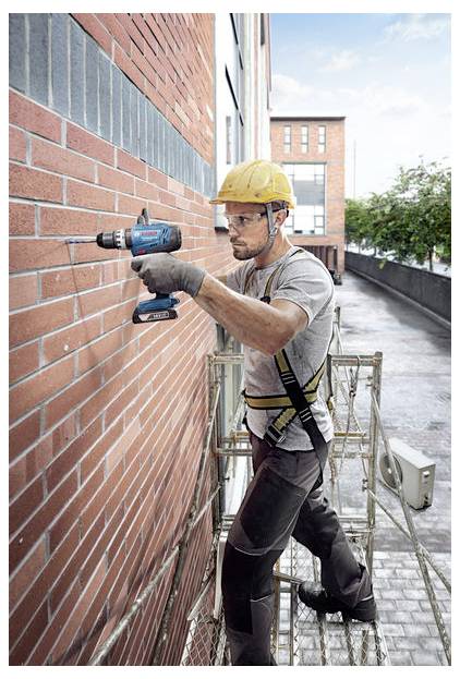 Ein Bauarbeiter mit Schutzhelm und Sicherheitsgurt benutzt einen Akkuschrauber an einer Ziegelwand, während er auf einem Gerüst außerhalb eines Gebäudes steht.
