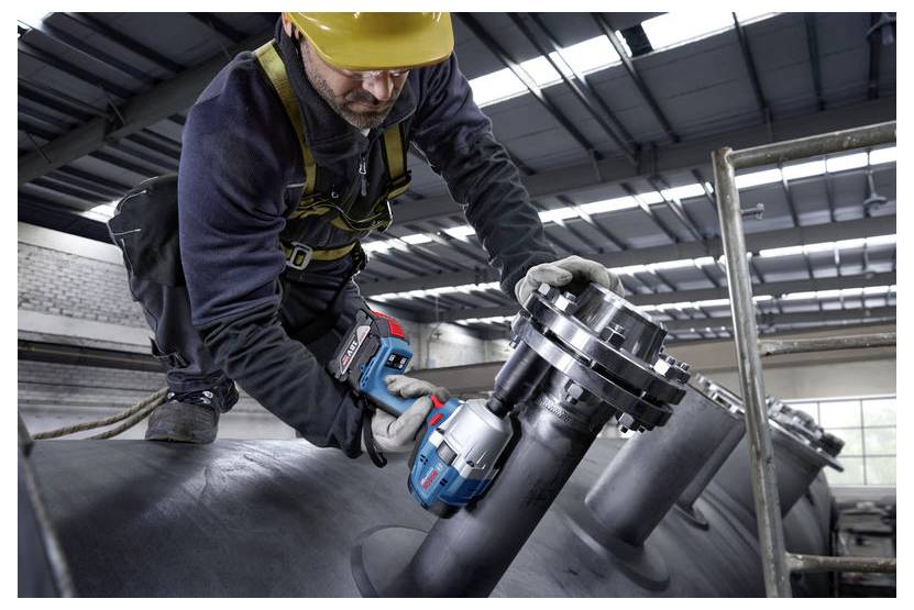 Ein Arbeiter mit Sicherheitsausrüstung und gelbem Schutzhelm verwendet ein Elektrowerkzeug, um Bolzen an einem großen Industrierohr in einer Fabrik anzuziehen.