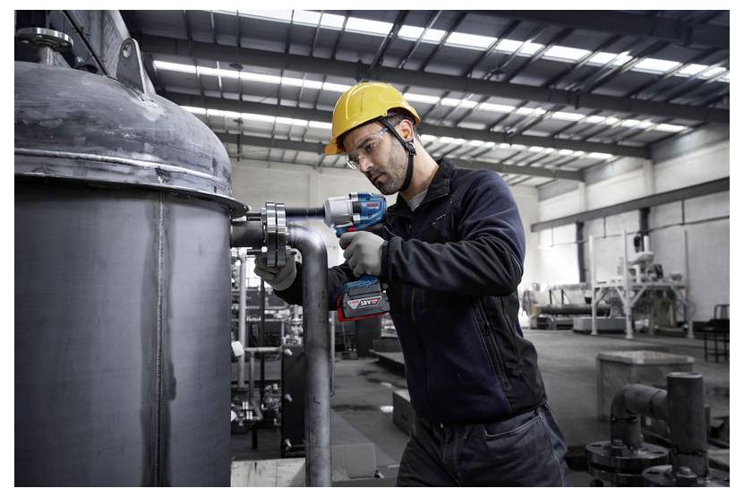 Ein Arbeiter in Sicherheitsausrüstung bedient ein Elektrowerkzeug an industriellen Rohrleitungen im Inneren einer großen Fabrik und unterstreicht dabei den Fokus auf industrielle Wartung.
