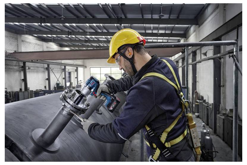 Ein Arbeiter in einem Sicherheitsgeschirr und Helm benutzt ein Elektrowerkzeug an einer großen Metallstruktur in einer Fabrikhalle.