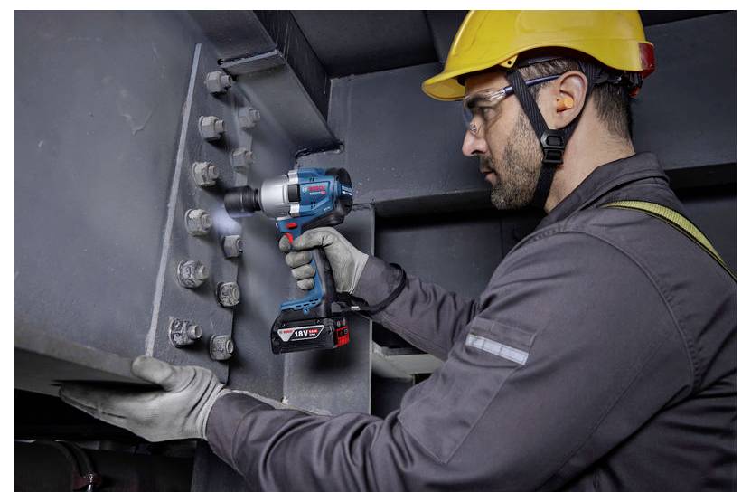 Ein Bauarbeiter mit Persönlicher Schutzausrüstung (PSA), einschließlich eines gelben Schutzhelms und Handschuhen, benutzt ein Elektrowerkzeug, um Schrauben an einer großen Metallkonstruktion zu befestigen.