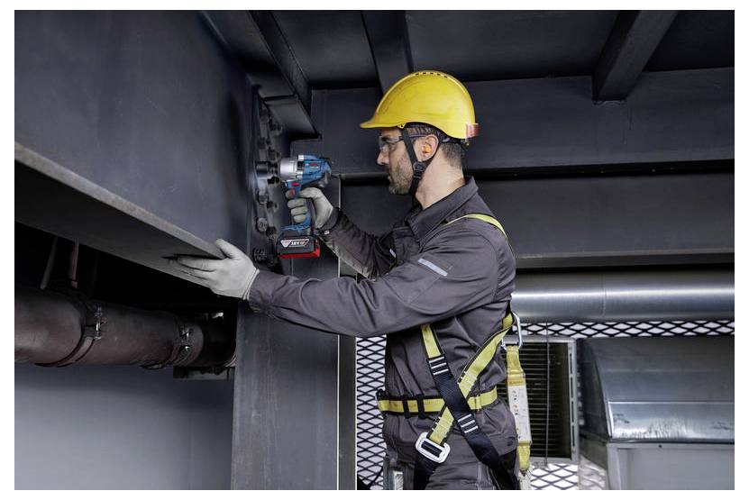 Ein Bauarbeiter in Sicherheitsausrüstung, einschließlich Helm und Sicherheitsgurt, verwendet ein Elektrowerkzeug, um Metallträger in einer industriellen Umgebung zu befestigen.