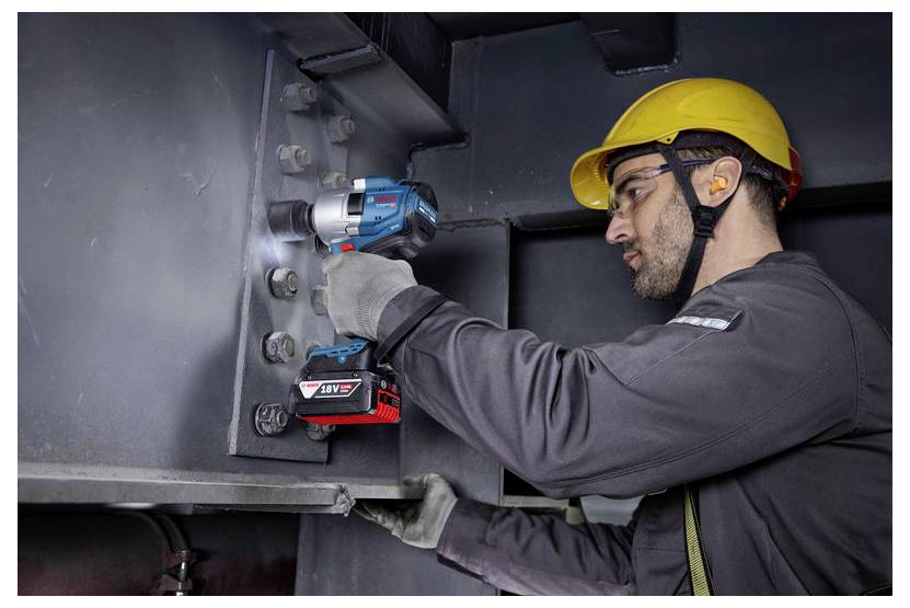 Ein Bauarbeiter mit einem gelben Helm benutzt ein blau-rotes Elektrowerkzeug, um Bolzen an einer Stahlkonstruktion zu befestigen.